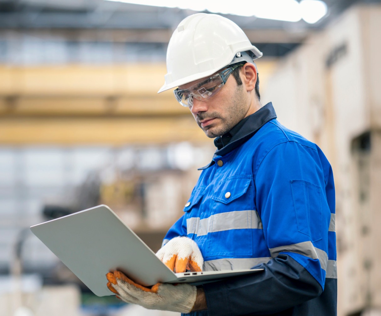 Factory Manager with hard hat and safety glasses viewing laptop on factory floor